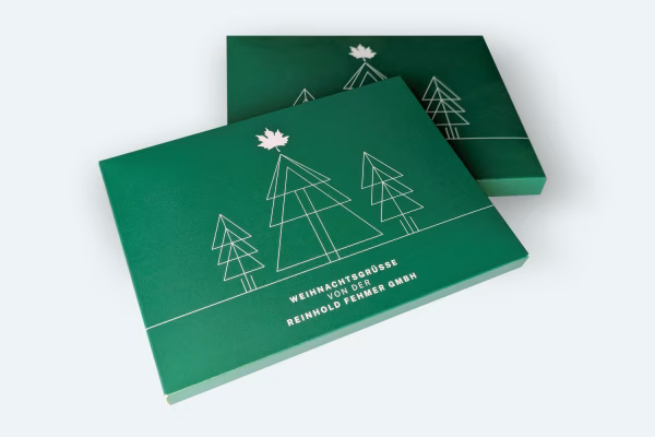Printdesign: Verpackung für Weihnachtspost Grüne Weihnachtskarten der Reinhold Fehmer GmbH mit minimalistischem Design: Geometrisch stilisierte Tannenbäume in weißen Linien und ein weißes Ahornblatt-Logo auf dunkelgrünem Hintergrund. Text 'Weihnachtsgrüße von der Reinhold Fehmer GmbH' in weiß. Zwei Karten sind leicht versetzt übereinander dargestellt.
