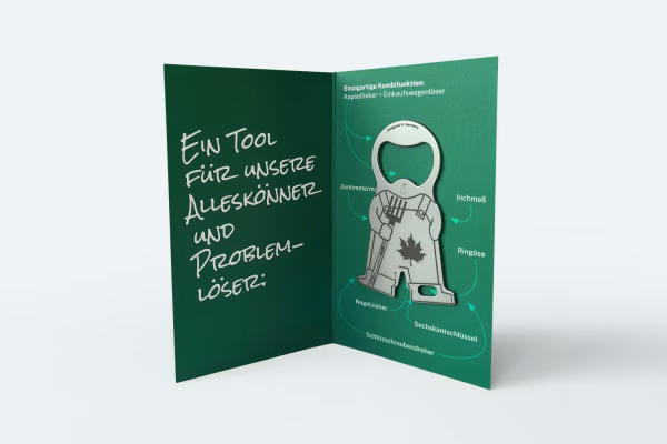 Printdesign: Give-Away Geöffnete grüne Karte mit einem silbernen Multifunktions-Werkzeug in Männchen-Form. Links handschriftlicher Text 'Ein Tool für unsere Alleskönner und Problemlöser.' Rechts Beschriftungen der verschiedenen Funktionen wie Kronkorkenöffner, Inchmaß, Ringöse, Sechskantschlüssel und Nagelzieher. Das Werkzeug ist mit dem Fehmer-Ahornblatt-Logo verziert.